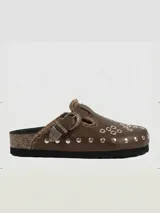 Zueco de cuero con diseño artesanal, presenta una tira con hebilla metálica decorativa, bordados sutiles y múltiples remaches metálicos en la parte frontal. El interior está forrado en cuero.