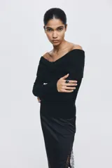 Buzo de punto acanalado negro con escote off-the-shoulder y detalle de nudo frontal. Presenta mangas largas y corte ajustado.