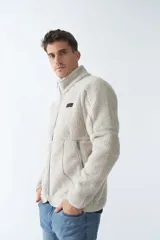 Campera de sherpa color beige con cierre frontal completo, cuello alto y bolsillos laterales con cierre. Presenta un parche decorativo en el pecho.