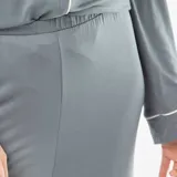 Pijama de dos piezas compuesto por una camisa de manga larga con botones frontales, cuello solapa y bolsillo en el pecho, y un pantalón de corte recto y holgado. Ambas prendas presentan un diseño liso en color gris con detalles de ribetes en contraste.