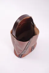Matera tipo bucket de cuero color bordó, con detalles de tiras verticales en cuero negro adornadas con remaches metálicos plateados. Cuenta con un asa de mano fija y un compartimento interior amplio.