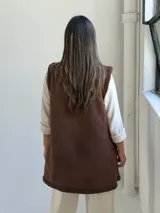 Chaleco largo de gamuza sintética color chocolate, con interior y solapas de corderito sintético al tono. Diseño abierto sin botones, con corte recto y largo por debajo de la cadera.