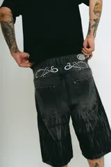 Bermuda de jean color negro con lavado acid wash gris. Presenta un corte baggy y detalles bordados en plateado y blanco que simulan ondas o llamas en la parte frontal superior e inferior.