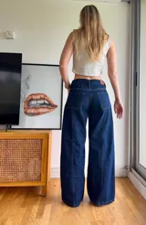 Jean de tiro medio en color azul índigo oscuro, con corte de pierna ancha y costuras verticales decorativas en el frente. Presenta bolsillos delanteros tipo parche y cierre de botón y cremallera.