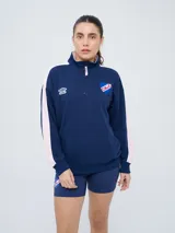 Buzo de felpa azul marino con medio cierre, cuello alto, bandas laterales en tono rosado y bolsillo frontal tipo canguro. Presenta el logo de Umbro y el escudo del Club Nacional de Football bordados.