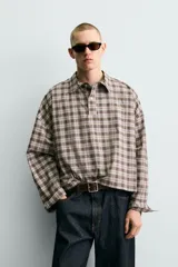 Camisa polo de corte boxy fit confeccionada en tejido de algodón con elasticidad, presenta un estampado de cuadros en tonos marrones, azules y blancos. Cuenta con cuello solapa, cierre frontal de botones, manga larga con puños abotonados y bajo con aberturas laterales.