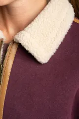 Campera tipo gamulán de color bordó, con interior y cuello de corderito sintético en tono crema. Presenta detalles de ribetes en cuero sintético color suela, cierre frontal metálico y bolsillos laterales.