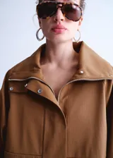 Chaqueta oversize color tostado con cuello embudo, manga larga, bolsillos laterales y bolsillos de parche con solapa. Cuenta con cierre delantero de cremallera y botones ocultos bajo tapeta, y forro interior.