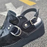 Zueco cerrado con plataforma, confeccionado en cuero legítimo negro con texturas combinadas de charol y gamuza. Cuenta con tira decorativa con argollas metálicas y tira trasera ajustable con hebilla.