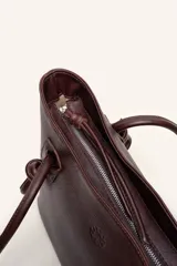 Cartera tote de cuero suave color marrón, con diseño amplio y líneas simples, con tiras de hombro decoradas con nudos artesanales y pequeño logo grabado en el centro.