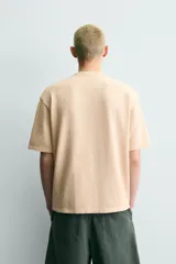Camiseta relaxed fit de punto color beige con efecto lavado, cuello redondo y manga corta. Cuenta con estampado frontal con texto en tonos amarillo, azul y rojo.