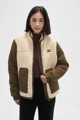 Campera de diseño híbrido que combina paneles de corderito sintético en color crema con mangas y bolsillos de tela técnica acolchada en color verde militar. Presenta cierre frontal completo, cuello alto y un pequeño parche decorativo en el pecho.