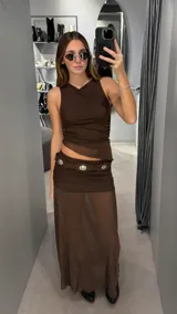 Conjunto de dos piezas color chocolate compuesto por un top sin mangas con cuello redondo y dobladillo asimétrico, y una pollera larga de tiro bajo con diseño fruncido en la cintura y falda transparente. Incluye un cinturón decorativo con apliques metálicos.