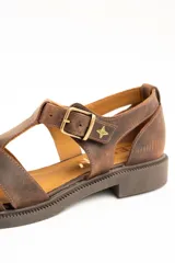 Sandalias tipo cangrejera de cuero color marrón, con diseño de tiras cruzadas, talón cerrado y tira ajustable con hebilla metálica y detalle de estrella grabada en el lateral. Suela plana con pequeño tacón.