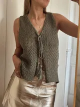 Chaleco tejido en punto calado con hilo de algodón y lurex, de color negro con destellos plateados. Presenta escote profundo en V, cierre frontal con cordones entrelazados terminados en borlas y un corte recto y holgado.