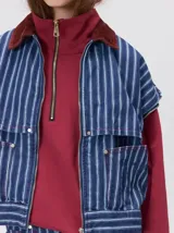 Campera de denim a rayas verticales con cuello de pana en color bordó. Presenta cierre frontal, mangas desmontables con cierre en los hombros, canesú en el pecho y bolsillos de parche cuadrados.