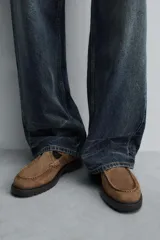 Zapato tipo Mary Jane de hombre, color marrón, fabricado en piel con acabado serraje (gamuza). Presenta cierre con hebilla lateral, detalle de bordón en el empeine y suela gruesa con volumen.