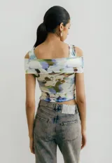 Top corto de jersey con estampado abstracto floral en tonos pastel (blanco, lila, verde, marrón). Presenta un diseño cruzado asimétrico en el busto que crea un escote tipo *off-shoulder* en un lado y manga corta en el otro.