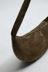 Bolso de hombro de piel con acabado serraje, color gris oscuro, con forma curva o de media luna. Presenta costuras visibles en el centro y un asa fina para llevar al hombro.