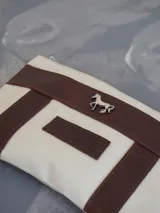 Sobre tipo cartuchera de lona naranja con detalles y apliques en cuero marrón. Presenta un aplique metálico en forma de caballo y un parche con el logo de la marca grabado en el frente.