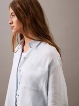 Camisa celeste de gasa doble, con cuello clásico, cierre frontal con botones, bolsillo de parche en el pecho y mangas largas con solapa enrollable.