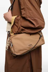 Bolso tipo morral Bimba y Lola, de color tostado, confeccionado en nylon suave con detalles en cuero y tirantes de cordón. Cuenta con múltiples compartimentos con cierre y el logo metálico de la marca en el frente.