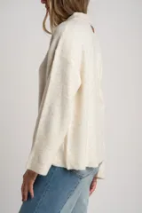 Sweater de tejido de punto color blanco roto o crema, con textura jaspeada. Presenta cuello polo y un escote con tres botones nacarados. El calce es holgado (oversize) y tiene mangas largas.