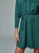 Vestido camisero color verde óxido, con cuello camisero, mangas largas con puños abotonados, dos bolsillos con solapa en el pecho, cinturón de lazo y cierre frontal con botones.