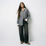 Camisa de corte oversize con diseño a rayas verticales en tonos verde y gris. Presenta cuello y puños en contraste de color blanco, cierre frontal con botones y un bolsillo en el pecho.