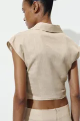 Camisa de lino con cuello solapa y escote en pico. Presenta un diseño sin mangas con detalle de pliegue cruzado en el delantero y cierre frontal con un botón.