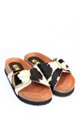 Sandalia tipo zueco con capellada de cuero con pelo estampado animal print de vaca en blanco y negro. Presenta una hebilla metálica dorada grande en el empeine, plantilla anatómica de corcho y suela de goma negra.