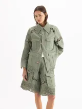 Chaqueta de gabardina liviana color verde militar, con silueta recta, largo por debajo de la cadera y cuello de corderoy. Presenta detalles de encaje en hombros, mangas y ruedo, múltiples bolsillos con solapa y cierre frontal con broches metálicos.