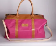 Bolso de viaje color fucsia con detalles en cuero color suela. Tiene doble asa de mano y correa larga desmontable.