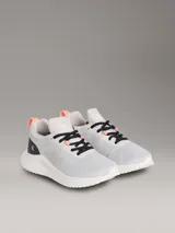 Zapatillas tipo runner Calvin Klein, de color gris con detalles en negro y coral. Presentan un diseño de malla perforada en la parte superior, cordones negros, suela chunky de EVA blanca y el logo de la marca en la lengüeta y el lateral.