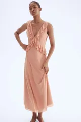 Vestido largo sin mangas de punto liviano, color rosado, con escote de pico pronunciado y ribetes de volado. Presenta volados de doble capa bajo el busto que se extienden por la cintura trasera. El vestido es forrado.