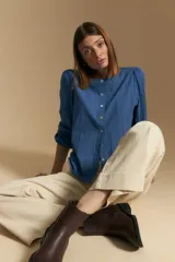 Camisa de algodón blanca con delicadas líneas bordadas verticales y textura calada. Presenta escote redondo con cierre frontal de botones, mangas largas con leve volumen en los hombros y puños elásticos.
