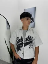 Camisa de corte boxy y manga corta, confeccionada en tela a cuadros gris y blanca. Presenta un diseño estampado frontal con tipografía estilo varsity, una estrella y detalles gráficos en negro.