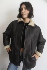 Campera de napalán de cuero genuino en color marrón oscuro, con interior y detalles en cuello y puños de piel sintética en tono crema. Presenta un diseño oversize con cierre frontal de botones y bolsillos laterales.