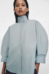 Camisa de algodón con cuello alto y mangas abullonadas por debajo del codo. Presenta cierre frontal con botones ocultos bajo solapa.