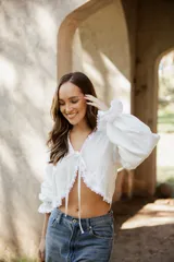 Blusa blanca de lino con mangas largas abullonadas, escote en V con lazo de ajuste frontal y detalles de puntilla en los bordes.