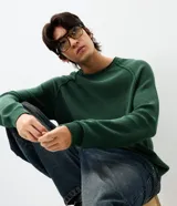 Suéter de punto color verde oscuro con textura, cuello redondo y mangas largas con diseño de corte raglán. Presenta puños y cintura acanalados, con un corte boxy moderno y estructurado.