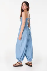 Mono largo de denim color celeste, sin mangas, con escote nido de abeja elástico y volado superior, tirantes finos para atar al cuello, perneras amplias de corte abombado con puños ajustables mediante lazos y bolsillos laterales.