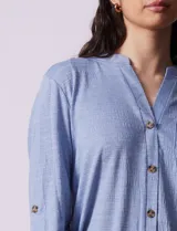 Blusa Ninety color beige con textura, cuello base con escote en V y abertura central con botones. Tiene un bolsillo en el pecho y mangas 3/4 regulables con presilla y botón.