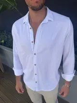 Camisa de manga larga confeccionada en lino, color blanco, con cuello clásico y cierre frontal de botones en tono madera.