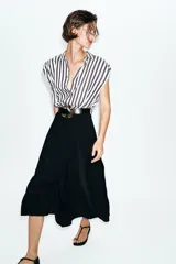 Vestido midi combinado con parte superior de camisa a rayas verticales blancas y negras, cuello solapa, manga sisa con frunces y botones dorados. La parte inferior es una falda midi negra de corte recto. Incluye un cinturón negro con textura de cocodrilo y hebilla dorada.