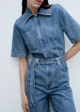 Mono largo de denim azul con cuello camisero, mangas cortas, cierre frontal con cremallera oculta, cinturón ajustable con hebilla metálica, bolsillos laterales y un bolsillo de parche en el pecho.