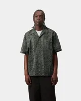 Camisa de manga corta con estampado de serpiente en tonos verdes y grises, cuello camisero abierto y cierre frontal con botones. Cuenta con un bolsillo en el pecho.