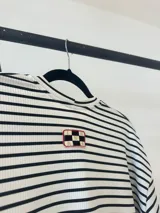 Remerón de manga larga con diseño de rayas horizontales finas en color negro sobre fondo blanco. Presenta un corte oversize, cuello redondo y un largo asimétrico con la parte trasera ligeramente más larga que la delantera.