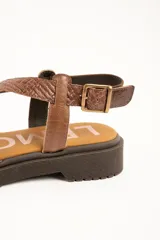 Sandalias de cuero color marrón con suela track, tiras cruzadas en el empeine con diferentes texturas y acabados, y tira ajustable al tobillo con hebilla metálica.