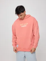 Canguro con capucha color salmón/coral desgastado, con efecto lavado. Presenta un estampado frontal con el texto "RUSTY & SUN" y un diseño de olas en color amarillo pálido. Tiene bolsillo canguro frontal y puños y cintura acanalados.